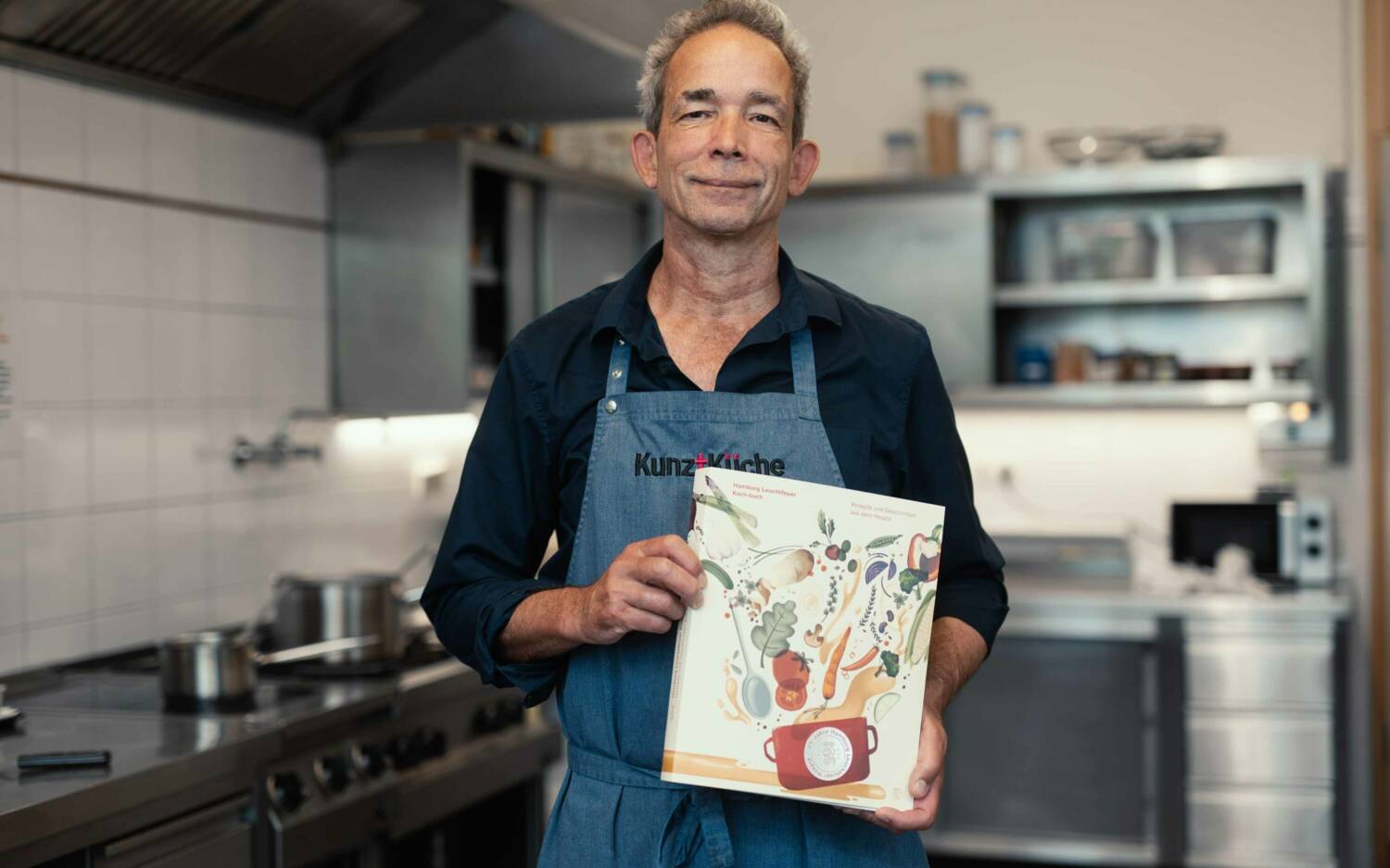 Küchenchef Ruprecht Schmidt hält das neue Kochbuch des Hamburg Leuchtfeuer Hospizes / ©Nico Vogelsaenger