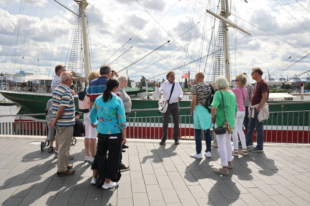 Eine Gruppe um einen Tourguide platziert vor einem angelegten Segelschiff.