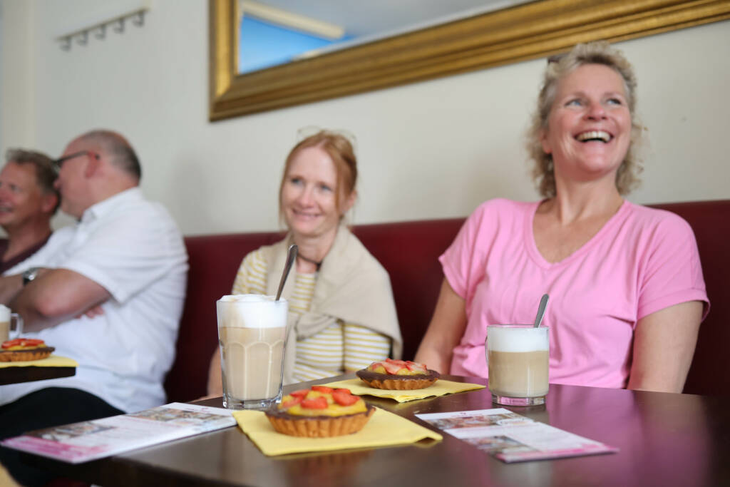 Zwei Damen in einem Cafe mit Heißgetränken und Törtchen. Beide lachen.