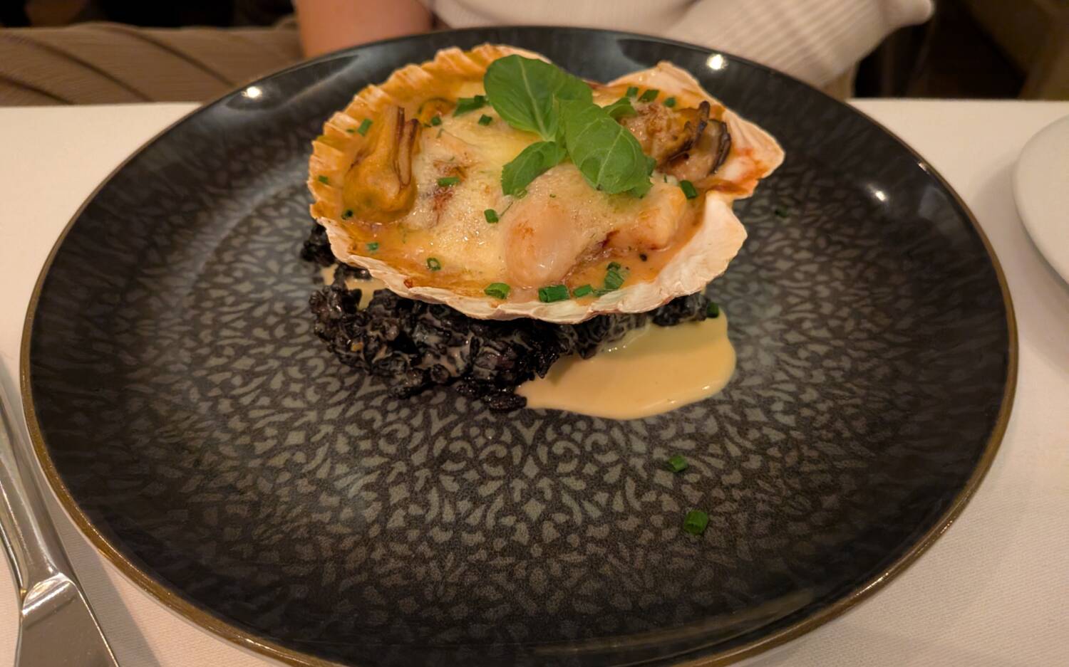 Spannender Einstieg im Charmant: Gratiniertes Muschelfleisch, präsentiert in einer Jakobsmuschel, die auf Sepia-Risotto liegt / ©Frédéric Guiot